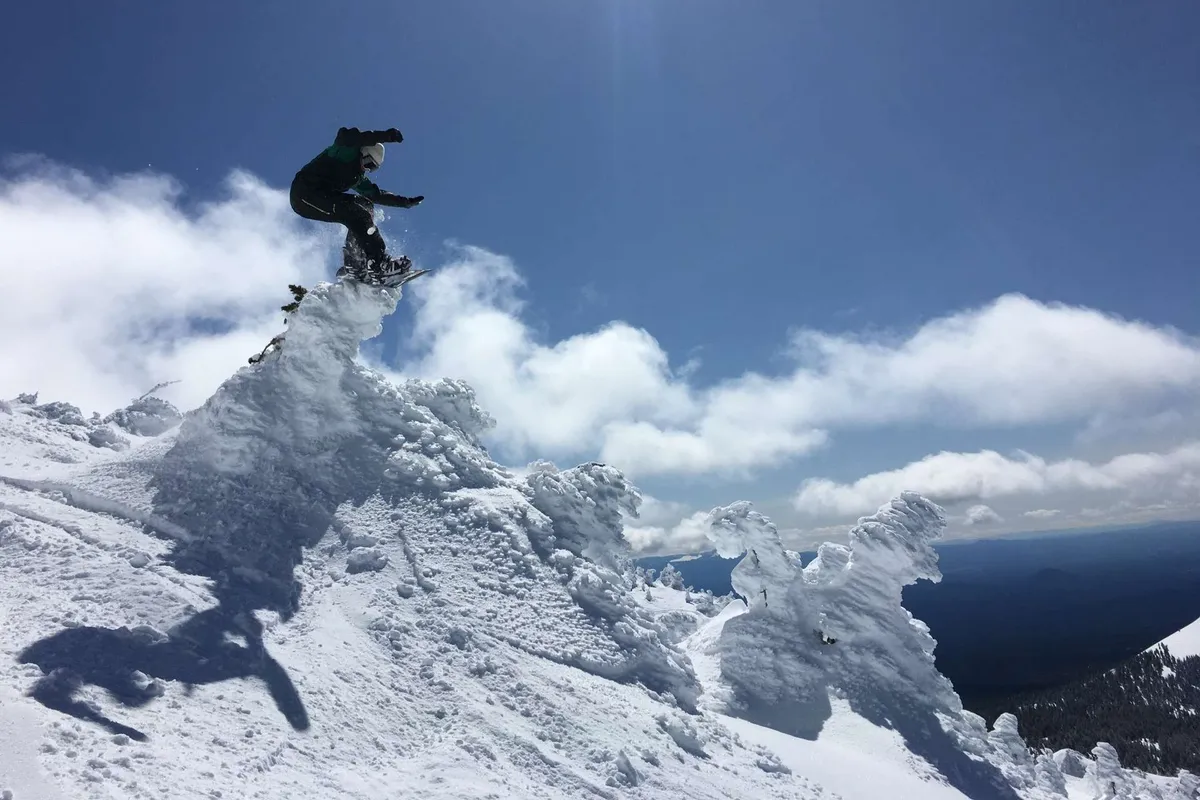 A dynamic action shot of a snowboarder in mid-air above a snow-covered mountain peak under a clear blue sky. The image conveys excitement, wintertime fun, and memorable experiences in the snow.