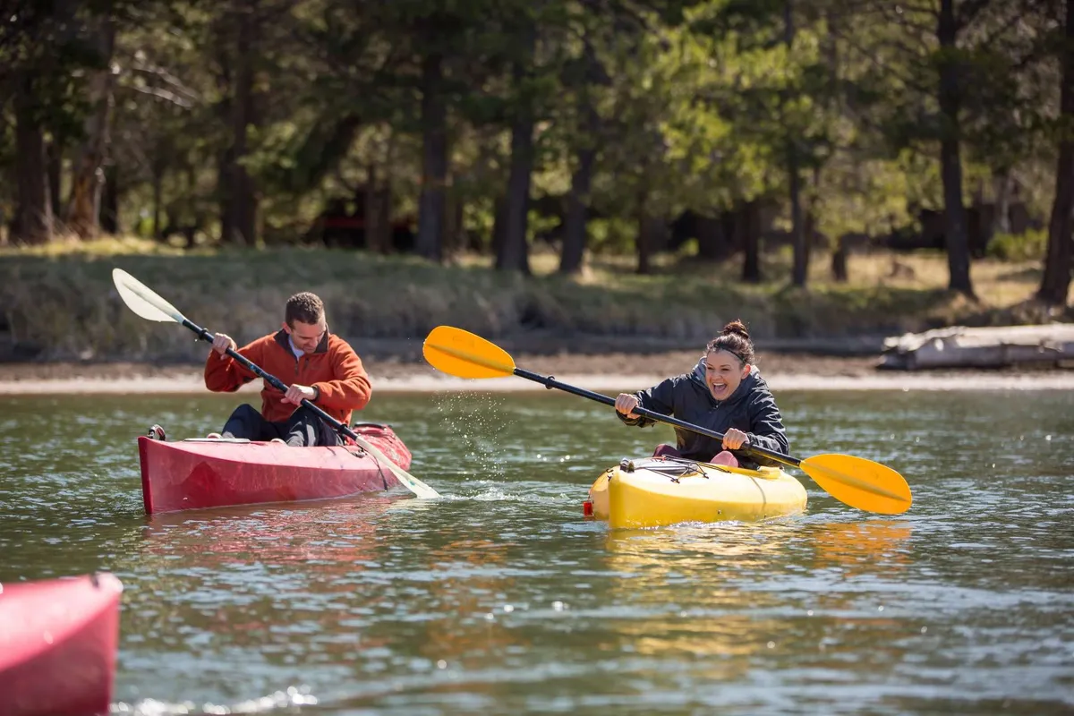 Two kayakers paddle through calm waters in bright, colorful kayaks, reflecting the joyful and adventurous spirit of water-based activities nearby. The natural surroundings suggest a vibrant outdoor context ideal for recreation.