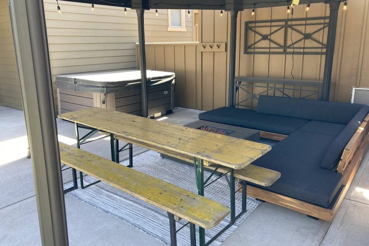 A wide exterior shot of a patio area featuring a dining table with outdoor seating and a hot tub nearby. The space is designed for social gatherings and relaxation, with the hot tub and dining area both visible, inviting guests to enjoy meals or unwind outdoors.