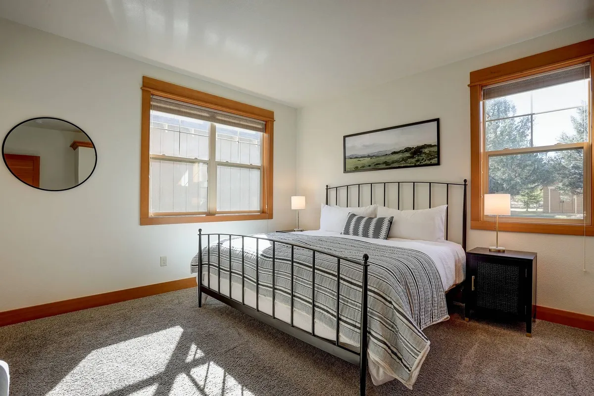 A serene bedroom featuring a bed with striped bedding, large windows letting in abundant natural light, a round mirror, and a nightstand with a lamp. The space feels both airy and cozy, ideal for unwinding after a long day.