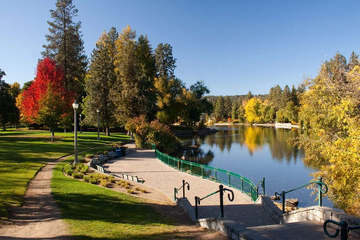 A tranquil lakeside scene with vibrant trees, a calm body of water, a walking path, and a distinctive green railing. The natural beauty of the landscape invites relaxed exploration and peaceful outdoor activities, such as walking, picnicking, or spotting local wildlife. The image conveys the wider resort environment accessible just steps from the Bunk House.