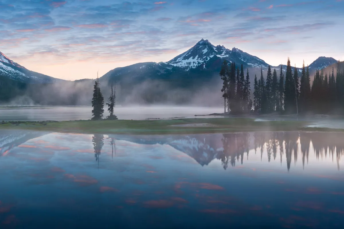 A breathtaking view of a tranquil mountain lake surrounded by lush trees and majestic peaks, with calm water reflecting the sky. The scene exudes natural beauty and offers a sense of peace and outdoor adventure, hinting at the scenic environment and recreational opportunities nearby.