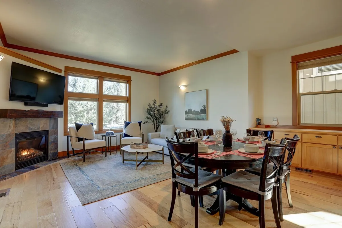 The image showcases a bright and welcoming living space featuring a cozy fireplace, a spacious dining table perfect for gatherings, a comfortable sofa area for relaxation, and large windows that fill the room with natural light. The setting is warm and inviting, ideal for family meals, fireside conversations, and unwinding after a day of resort adventures.