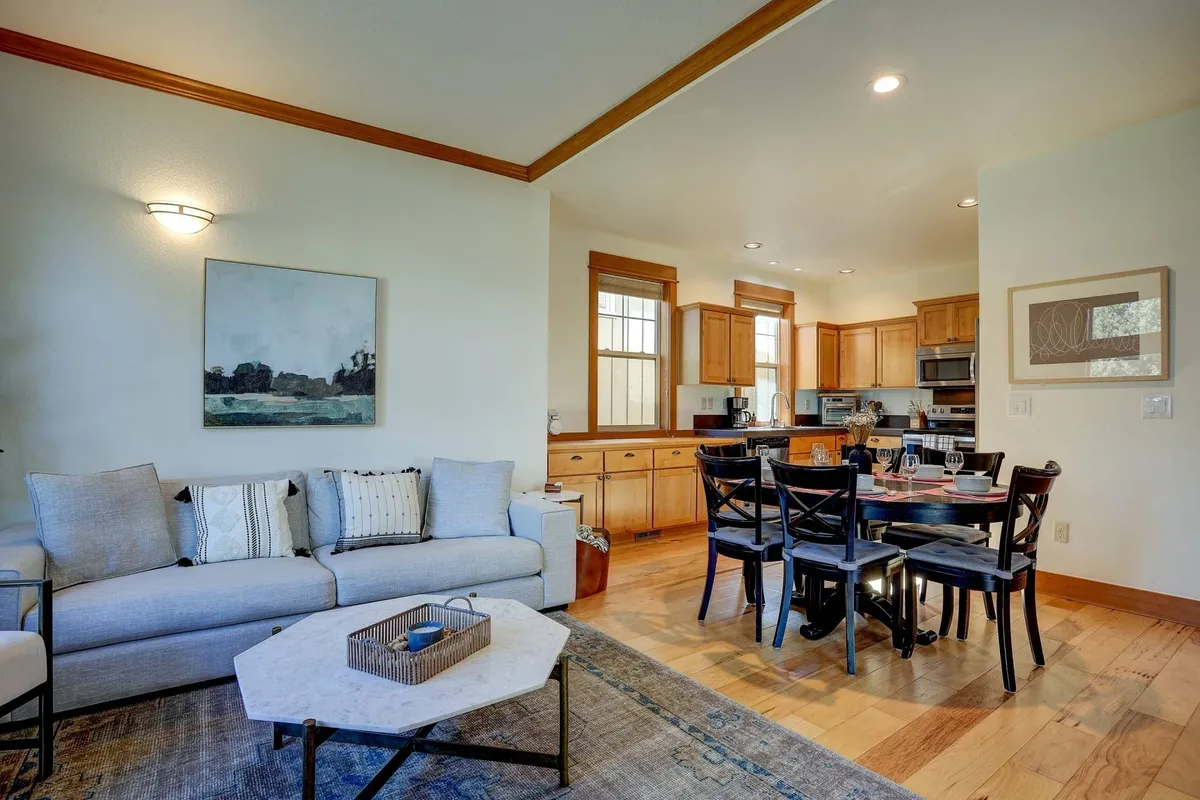 An inviting open-concept space featuring a cozy sofa, a view of the fully equipped kitchen, a spacious dining table set for six, and vibrant artwork on the wall. The room exudes warmth and comfort, ideal for gatherings and shared meals.