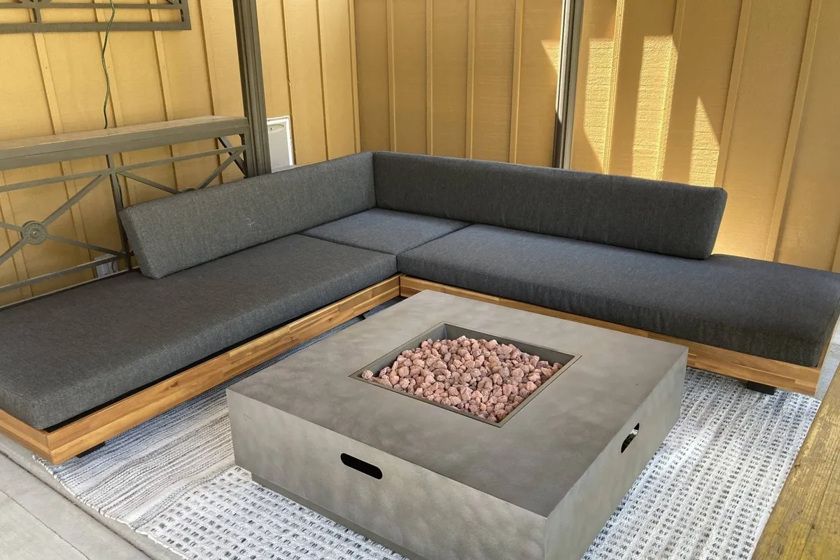A cozy sectional couch surrounds a fire pit atop a textured rug, creating a welcoming space for socializing, relaxing, or enjoying snacks together. The setup evokes the warmth of shared evenings, whether roasting s'mores, chatting with friends, or enjoying a drink under the stars. The design bridges indoor comfort with outdoor ambiance.