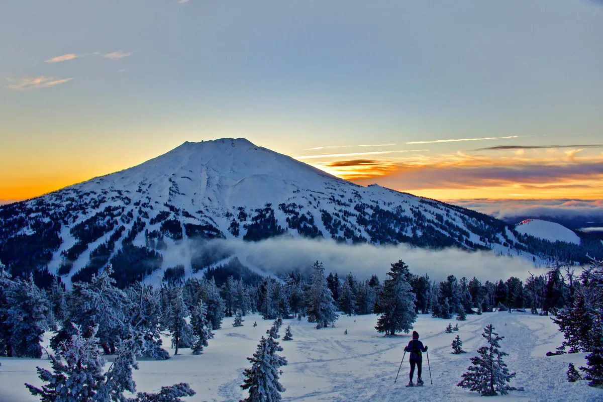 A dramatic winter landscape with a snow-covered mountain, a skier in motion, and trees dusted in white. The sun sets behind the peaks, creating a sense of exhilaration and natural beauty.